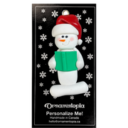 Ornament of white snowman in red hat holding a green book, on display backing card.