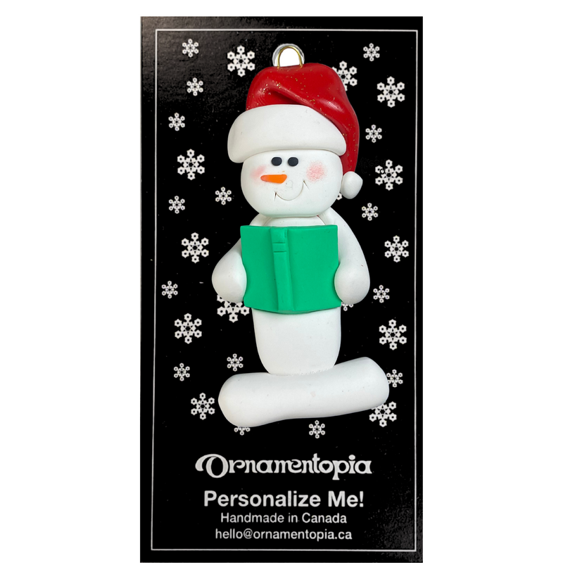 Ornament of white snowman in red hat holding a green book, on display backing card.