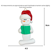 Ornament of white snowman in red hat holding a green book, graphed to show size (4.5x9cm).