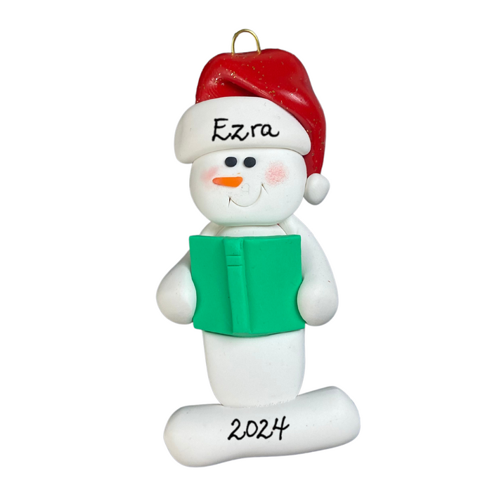 Ornament of white snowman in red hat holding a green book, personalized with name.