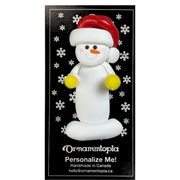 Ornament of white snowman in red hat holding yellow cheerleader pom-poms, on display backing card.