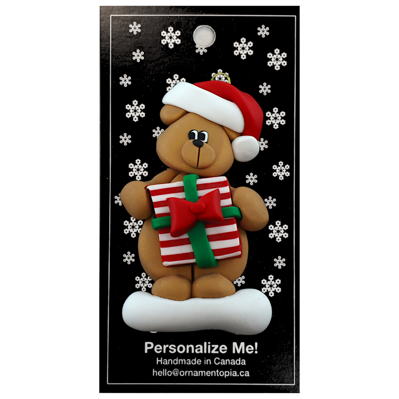 Tan bear in red festive hat holding colourful present ornament, on display backing card.