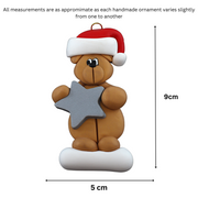Tan bear in red festive hat holding silver star ornament, graphed to show size (5x9cm).