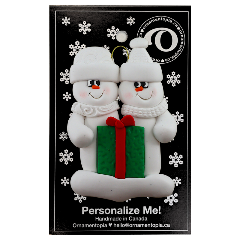 A couple’s ornament of two white snowmen holding a vibrant present, on display backing card.