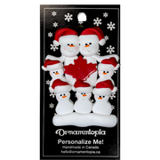 Family ornament with 7 white snowmen in red hats, gathered around a red maple leaf, on display backing card.