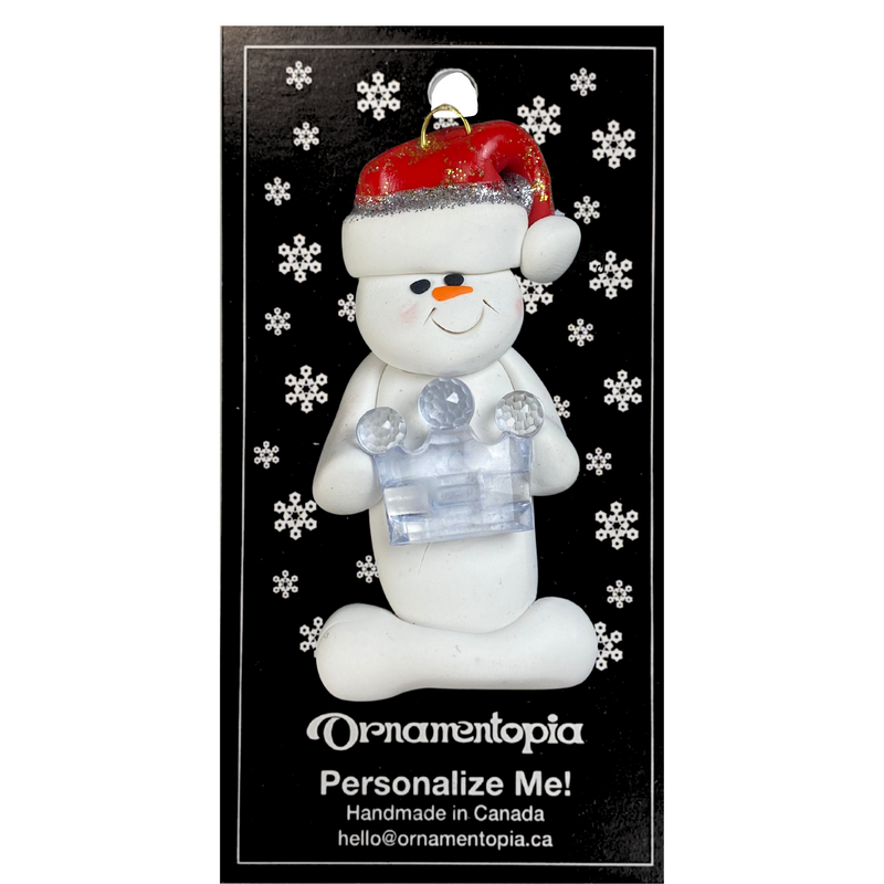Ornament of white snowman in red hat holding clear plastic crown, on display backing card.