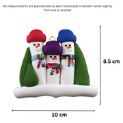 Family ornament with 3 snowmen in colourful scarves and hats, hemmed by Christmas trees, graphed to show size (10x8.5cm).