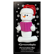 Ornament of white snowman in red hat holding a pink book, on display backing card.