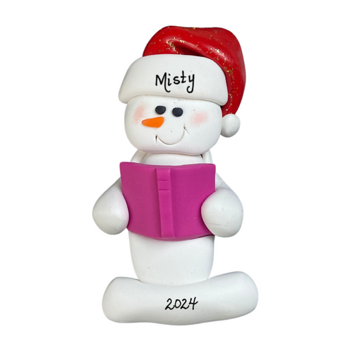 Ornament of white snowman in red hat holding a pink book, personalized with name.