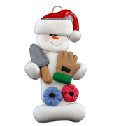 Ornament of white snowman in red hat holding a gardening glove and a shovel with flowers.
