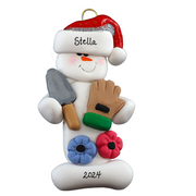 Ornament of white snowman in red hat holding a gardening glove and a shovel with flowers, personalized with name.