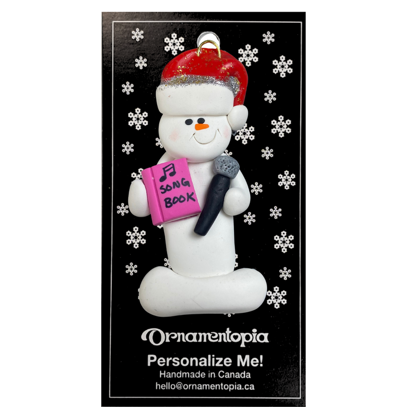 Ornament of white snowman in red hat holding a microphone and pink music book, on display backing card.