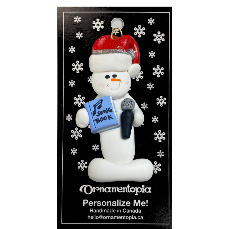 Ornament of white snowman in red hat holding a microphone and blue music book, on display backing card.