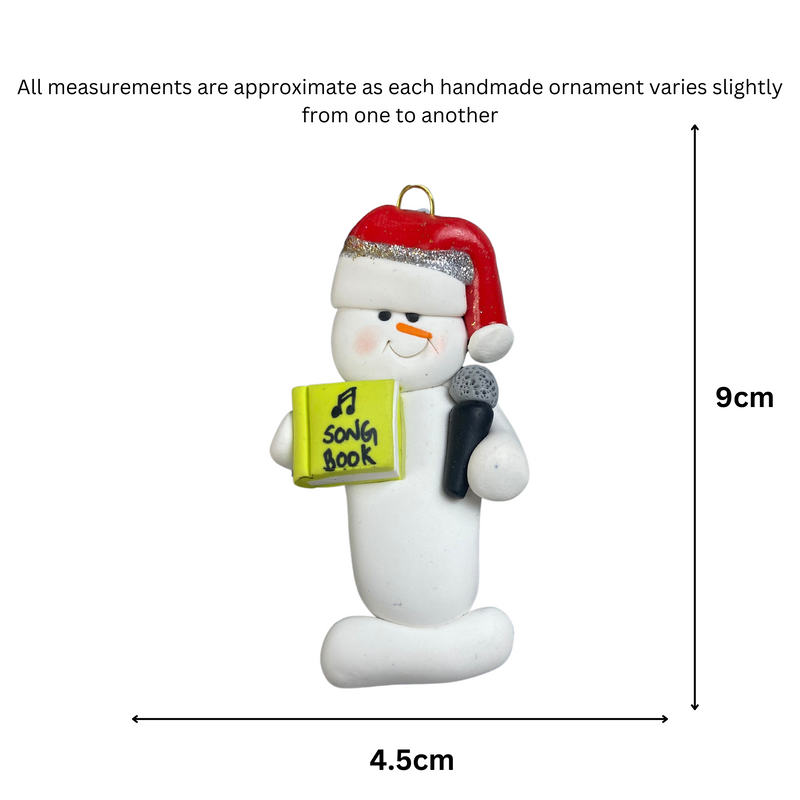 Ornament of white snowman in red hat holding a microphone and green music book, graphed to show size (4.5x9cm).