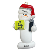 Ornament of white snowman in red hat holding a microphone and green music book, personalized with name.