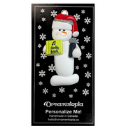 Ornament of white snowman in red hat holding a microphone and green music book, on display backing card.