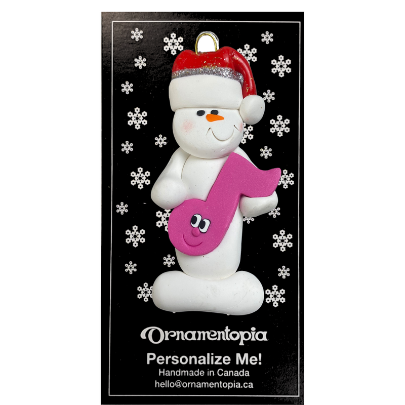 Ornament of white snowman in red hat holding a pink music note, on display backing card.