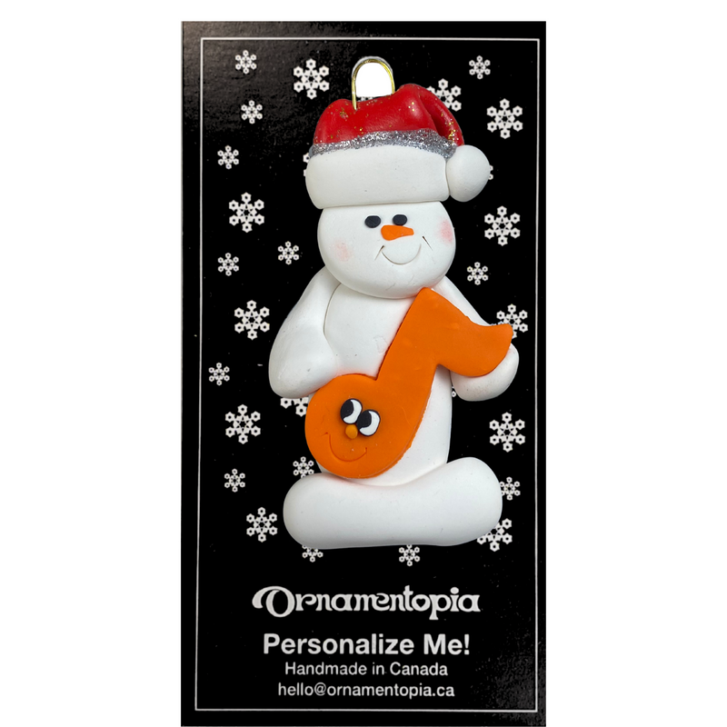 Ornament of white snowman in red hat holding an orange music note, on display backing card.