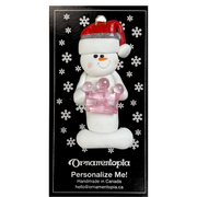 Ornament of white snowman in red hat holding a pink plastic crown, on display backing card.