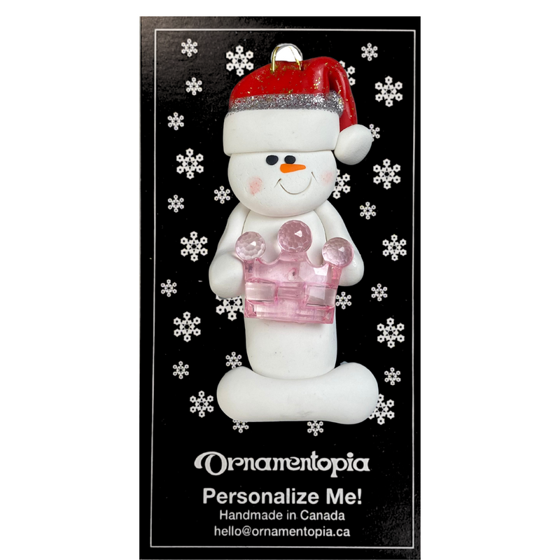 Ornament of white snowman in red hat holding a pink plastic crown, on display backing card.