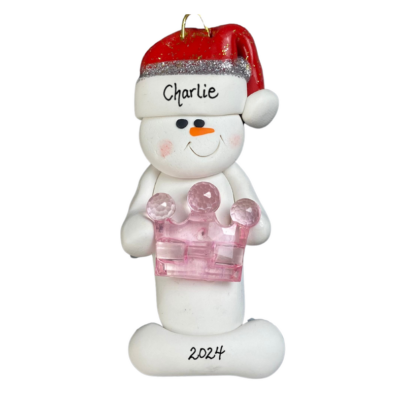 Ornament of white snowman in red hat holding a pink plastic crown, personalized with name.