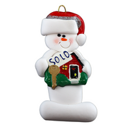 Ornament of white snowman in red hat holding a sign with a house saying “sold”.