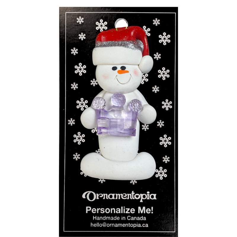 Ornament of white snowman in red hat holding purple plastic crown, on display backing card.