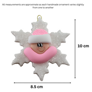 Baby ornament with infant face and pink hat on glittery white snowflake base, graphed to show size (8.5x10cm).