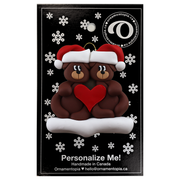 A couple’s ornament of two brown bears holding a red heart together, on display backing card.