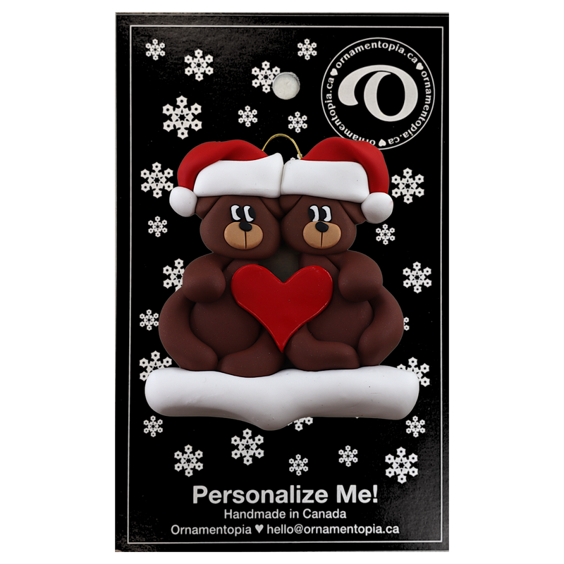 A couple’s ornament of two brown bears holding a red heart together, on display backing card.