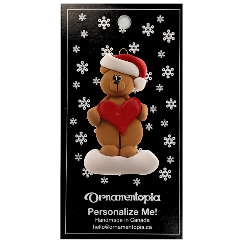 Tan bear in red festive hat holding red heart ornament, on display backing card.