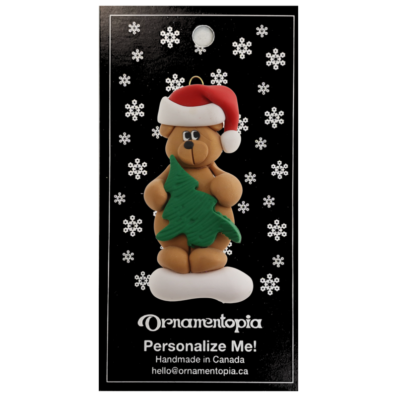 Tan bear in red festive hat holding green tree ornament, on display backing card.