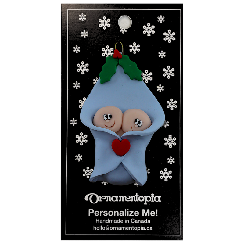 Ornament with twin infant heads poking out of a blue baby blanket, on display backing card.