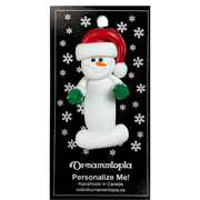 Ornament of white snowman in red hat holding green cheerleader pom-poms, on display backing card.