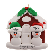 A couple’s ornament of two small white snowmen beside a festive brown house, personalized with names.