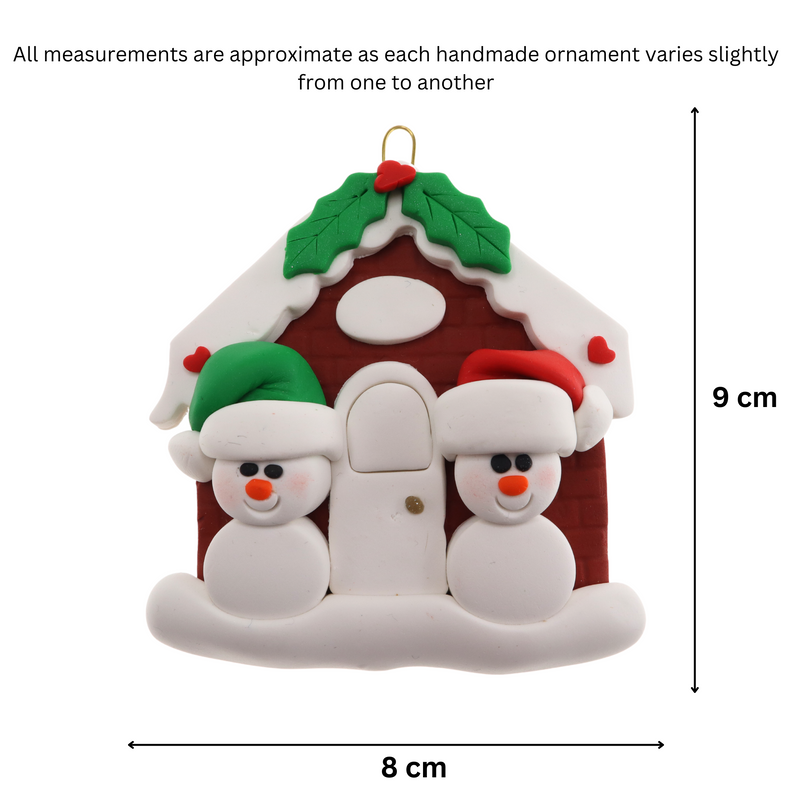 A couple’s ornament of two small white snowmen beside a festive brown house, graphed to show size (8x9cm).