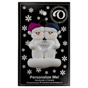A couple’s ornament of two white snowmen holding a plastic diamond, on display backing card.
