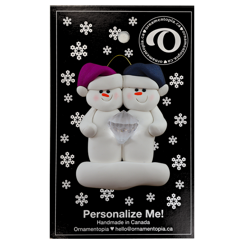 A couple’s ornament of two white snowmen holding a plastic diamond, on display backing card.