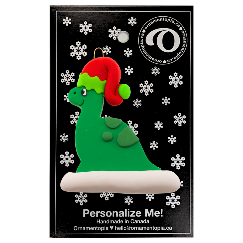 Green dinosaur wearing red festive hat ornament, on display backing card.