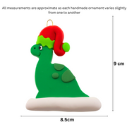 Green dinosaur wearing red festive hat ornament, graphed to show size (8.5x9cm).
