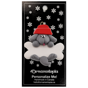 Ornament of grey dog in red hat hanging on a white bone, on display backing card.