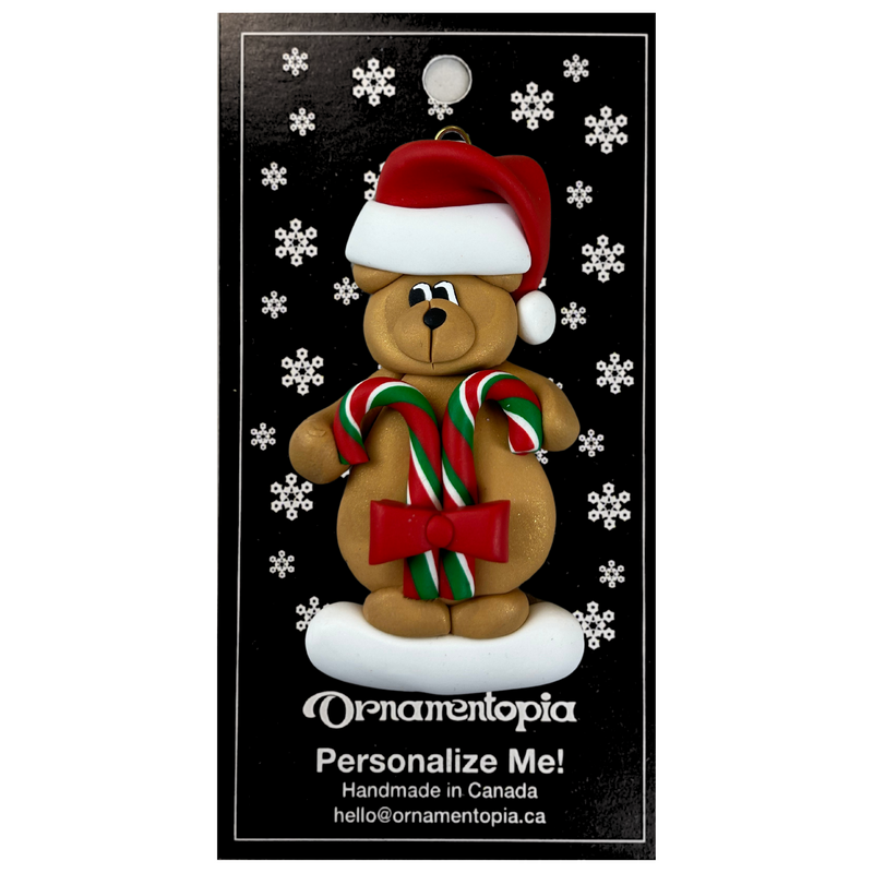 Tan bear in red festive hat holding two candy canes ornament, on display backing card.
