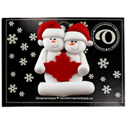 Family ornament with 2 white snowmen in red hats, gathered around a red maple leaf, on display backing card.