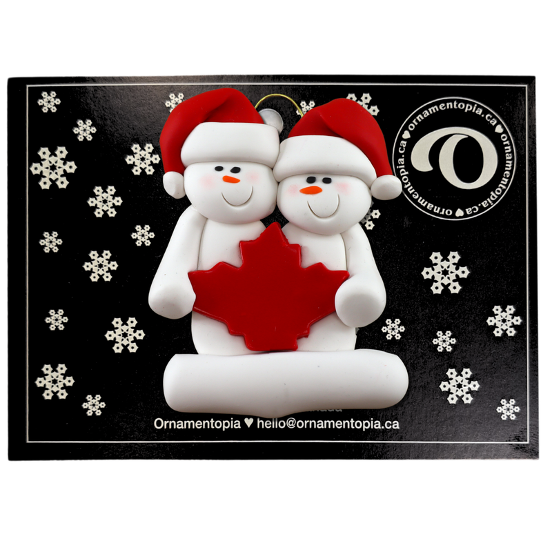 Family ornament with 2 white snowmen in red hats, gathered around a red maple leaf, on display backing card.
