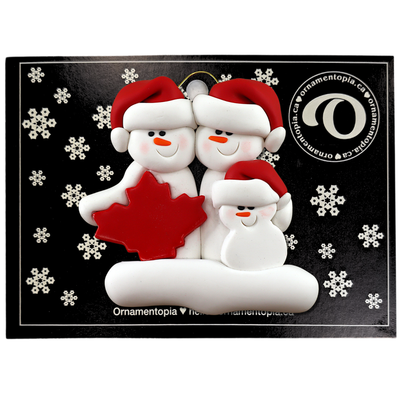 Family ornament with 3 white snowmen in red hats, gathered around a red maple leaf, on display backing card.