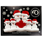 Family ornament with 6 white snowmen in red hats, gathered around a red maple leaf, on display backing card.
