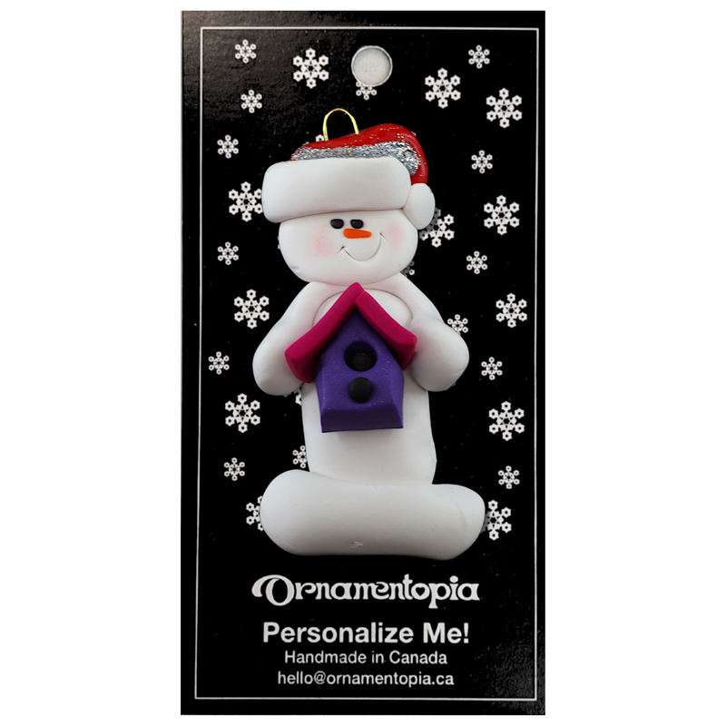 Ornament of white snowman in red hat holding a pink and purple birdhouse, on display backing card.