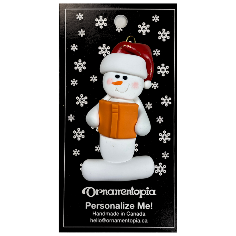 Ornament of white snowman in red hat holding an orange book, on display backing card.