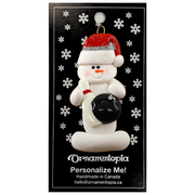 Ornament of white snowman in red hat holding a black bowling ball and pin, on display backing card.