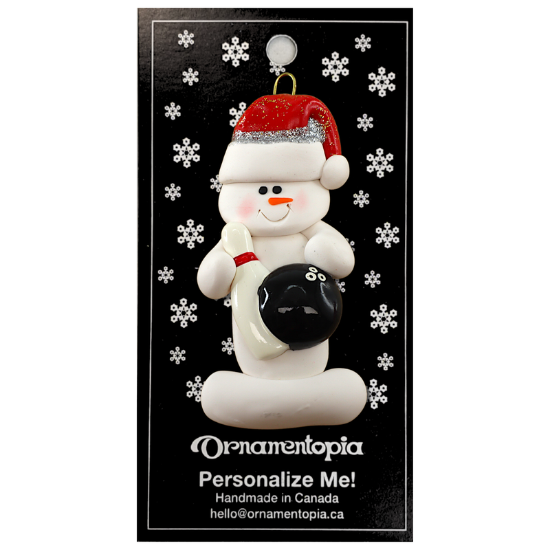 Ornament of white snowman in red hat holding a black bowling ball and pin, on display backing card.
